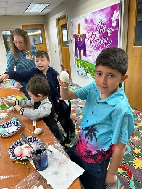 Children decorating Easter eggs at a table during an activity, with a woman assisting in the background. The room has a colorful carpet and a pink sign on the wall that says 'He is Risen.'
