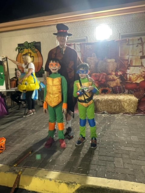 Two children dressed as Teenage Mutant Ninja Turtles standing with an adult in a cowboy hat at a Halloween event, with a Halloween-themed backdrop and other children in costumes in the background.