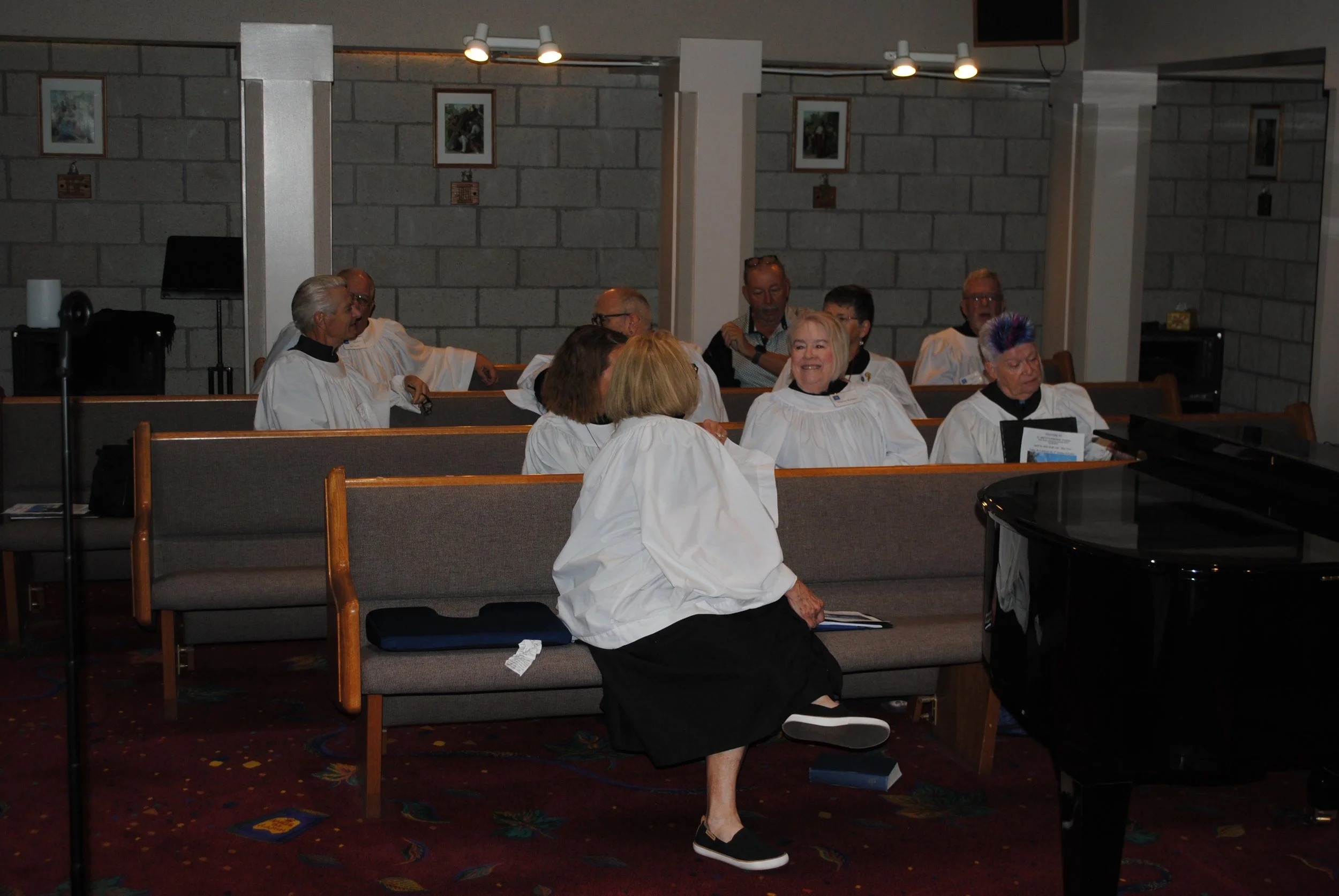 A group of elderly people wearing white choir robes sitting on benches inside a church, engaged in conversation and smiling.