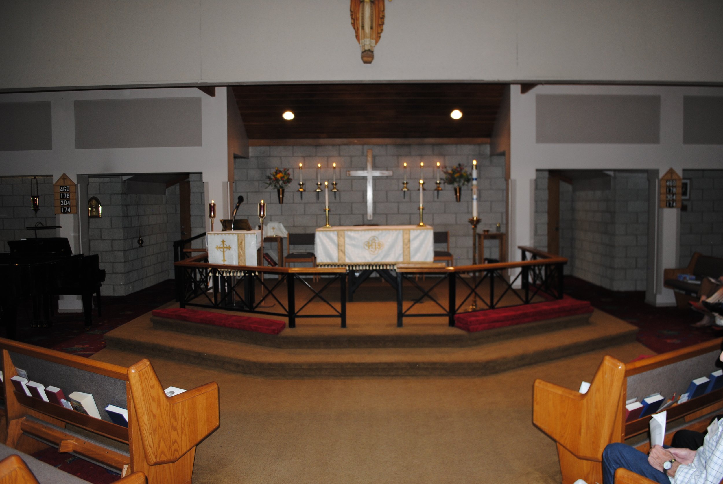 Inside a church, view of the altar with a white cross, lit candles, floral arrangements, and hymnals in wooden pews.
