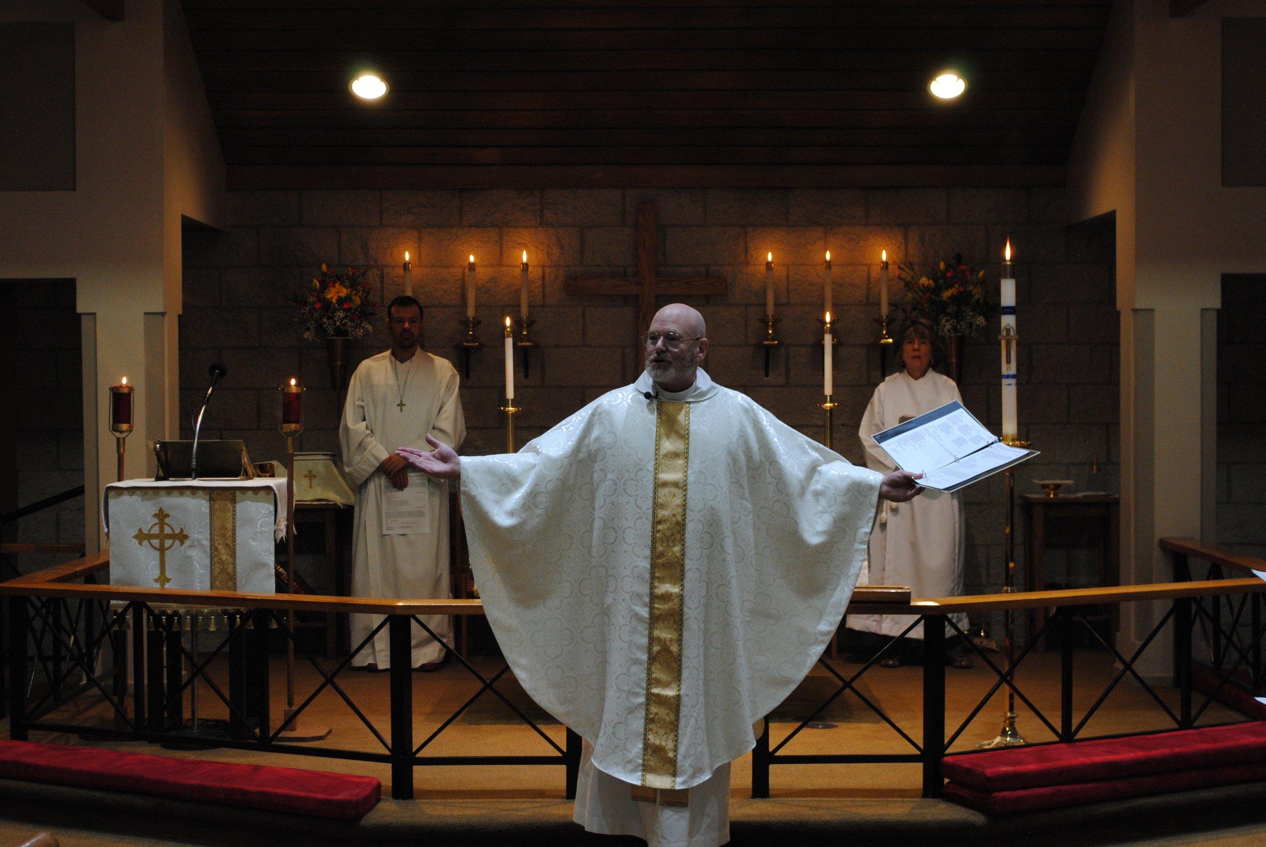 A priest conducting a ceremony in a church, with two assistants standing behind on either side. The priest is wearing white and gold vestments, with arms outstretched. The altar is decorated with candles, flowers, and religious symbols, and a wooden 