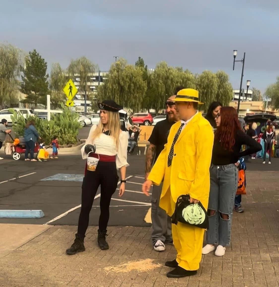 A group of people dressed in costumes standing and chatting in a parking lot during daytime, with children and other people in the background.