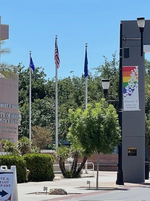 Three flagpoles with flags, trees and a rainbow pride flag on a building, street lamps, and a sidewalk in a city park setting during the daytime.