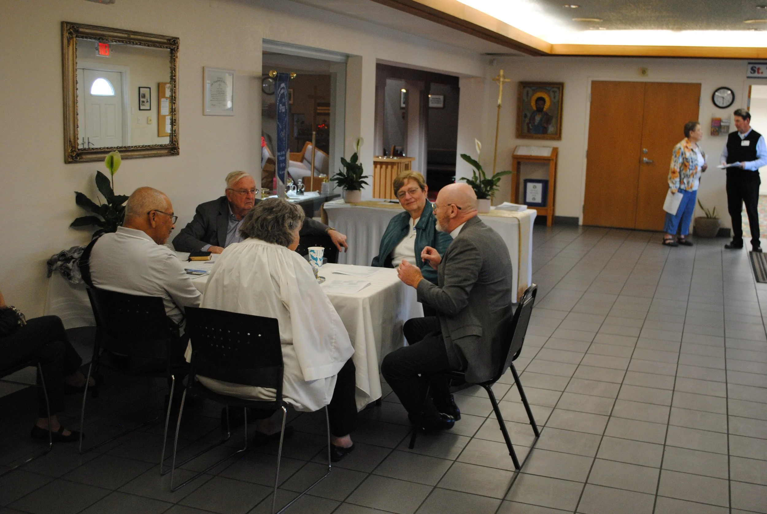 A group of five people sitting at a round table in a room, engaged in conversation. Two people are standing near a door in the background, smiling and talking. The room has religious pictures on the wall and a large mirror, with potted plants and inf