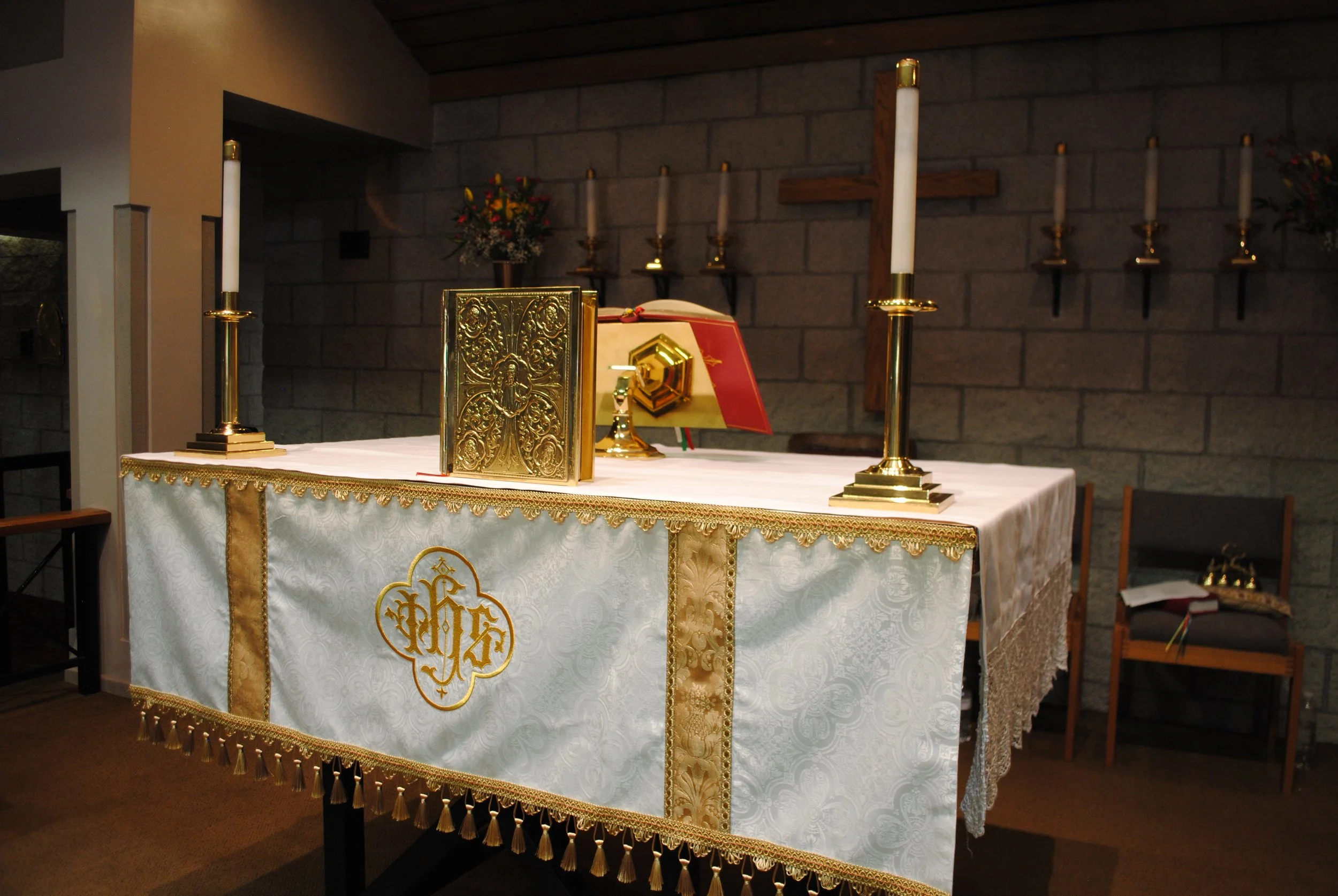 Inside a church or chapel, an altar with a white cloth featuring gold embroidery and tassels. On the altar, there are two tall white candles with gold holders, a golden ornate book cover, an open liturgical book, and a chalice. A wooden cross is moun
