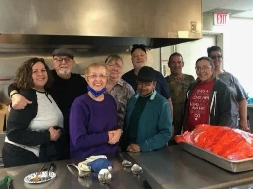 A group of nine diverse people, including children and adults, gathered together in a kitchen or food service area. They are smiling and standing around a counter with a large tray of prepared food covered with plastic wrap.