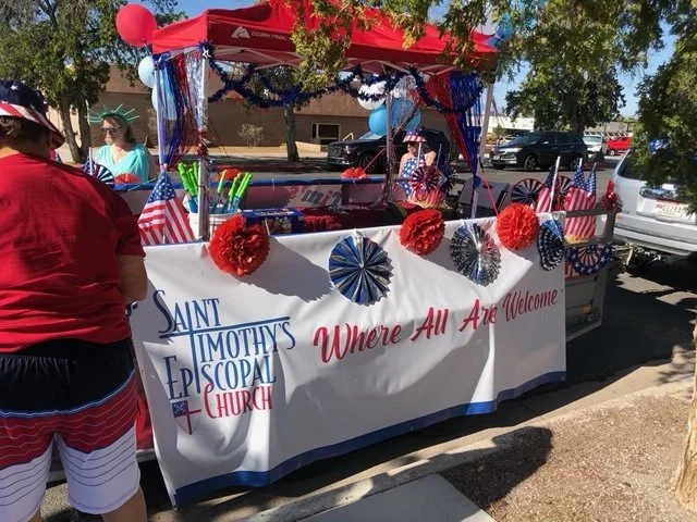 A decorated float for a local parade, featuring a banner for Saint Timothy Episcopal Church with the message "Where All Are Welcome," adorned with red, white, and blue decorations, including paper fans, balloons, and garlands. Several people are arou