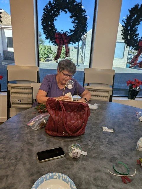 An elderly woman sitting at a conference table, reading a book, with holiday decorations including wreaths and poinsettias in the background.