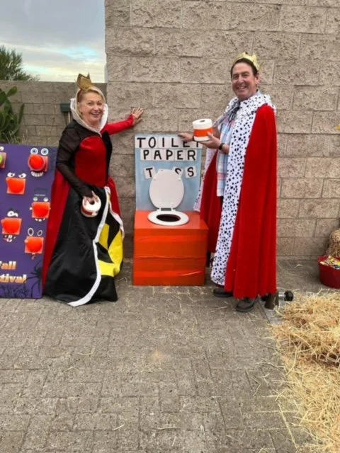 Two people dressed as kings or royalty standing next to a cardboard cutout of a toilet with a sign that says "Toilet Paper TOSS". They are smiling and holding toilet paper rolls, participating in a fun outdoor event.