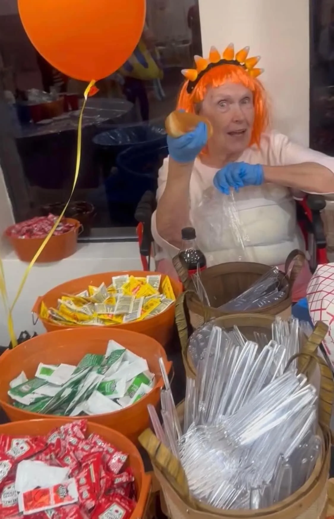 Woman with orange wig and costume holding a hot dog at a festive gathering with buckets of candy and orange balloons.