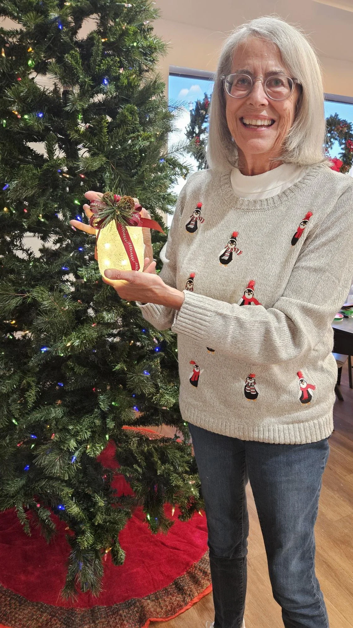 Senior woman celebrating Christmas, smiling, holding a lit decorative lantern with a red bow and pine cone, standing next to a decorated Christmas tree.