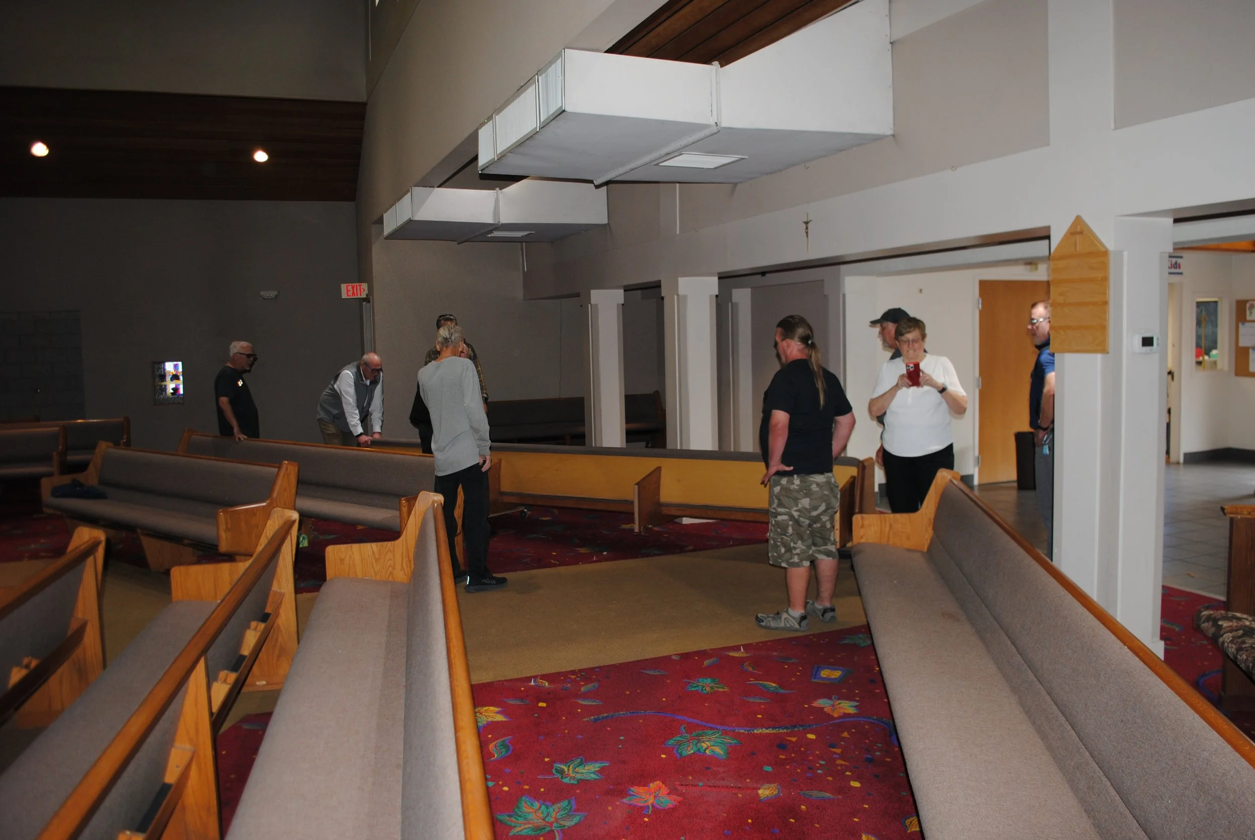Interior of a church or community hall with empty pews and several people standing and talking.