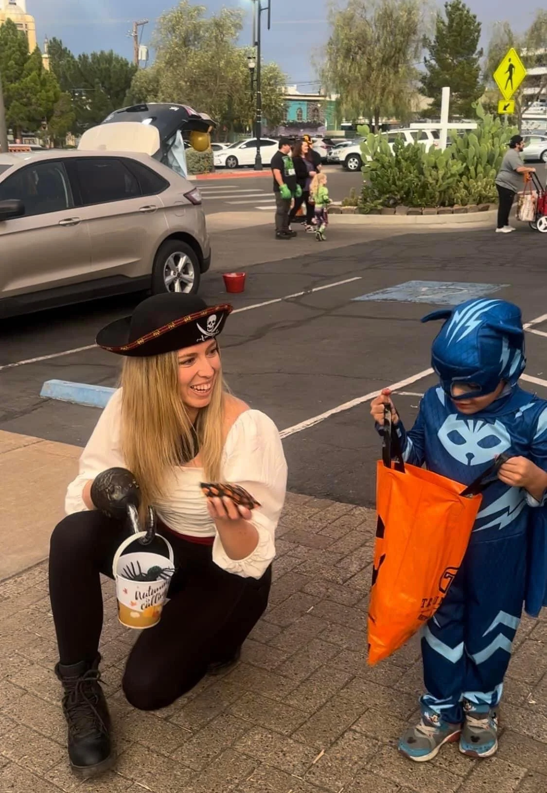 A woman dressed as a pirate kneeling on the sidewalk, smiling, talking to a young boy in a superhero costume. The woman holds a container of cold brew coffee with the signature 'Autumn Coffee' logo, and the boy holds an orange pumpkin-shaped trick-or