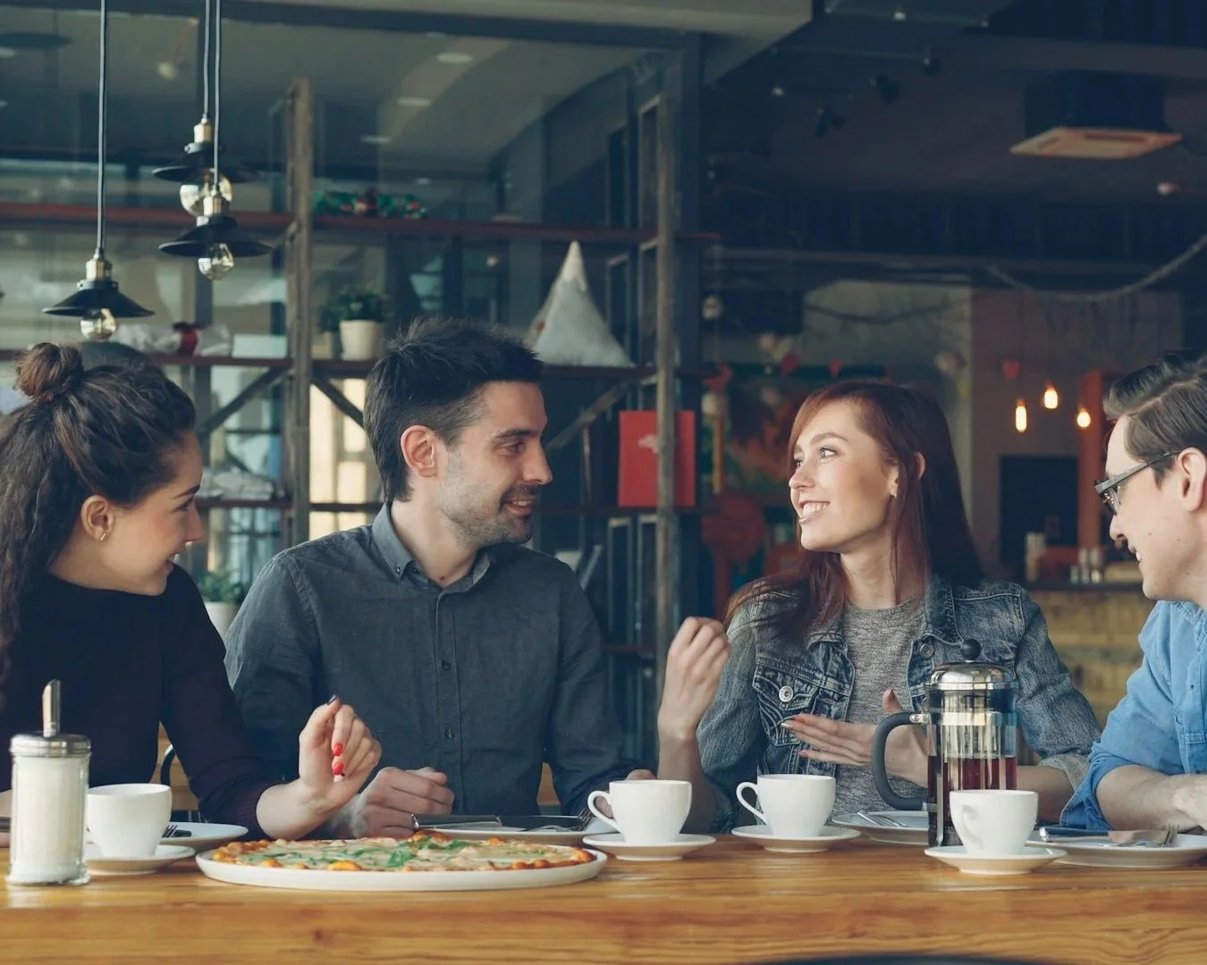 A group of five young adults sitting at a wooden table in a cafe, engaged in conversation with coffee cups, a pizza, and a French press on the table.
