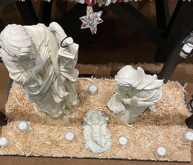 Nativity scene with white figurines of Mary, Joseph, and baby Jesus on a bed of straw, surrounded by small white candles, and a star hanging above.