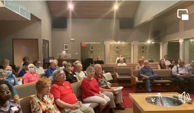 A group of people seated in a church or community hall, facing forward, listening to a speaker or presentation. The room has wood-paneled ceiling and pews with some people wearing masks.