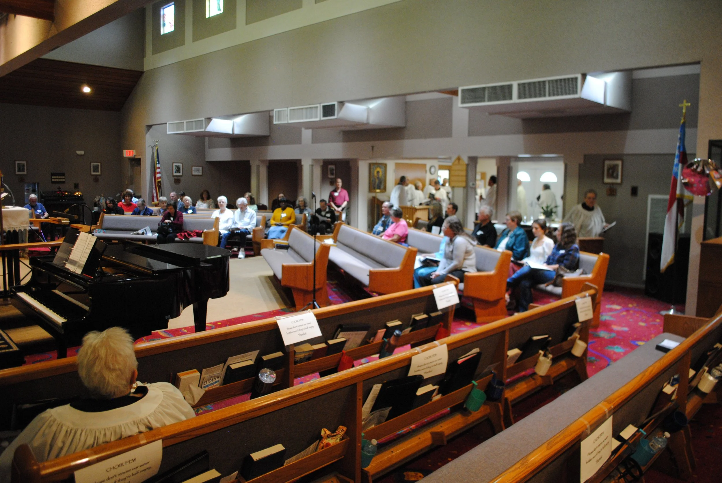 A group of people seated in pews inside a church, listening to a speaker or choir. A piano is in the foreground, and flags are positioned at the front. The church has high ceilings, stained glass windows, and religious artwork.