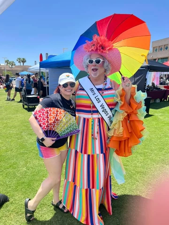 A woman dressed in a colorful, striped dress with a sash that reads 'Mrs Las Vegas Pride' is standing next to a person in a rainbow-colored outfit, both smiling. The woman is holding a rainbow fan and has a large, rainbow-colored umbrella, with a lar