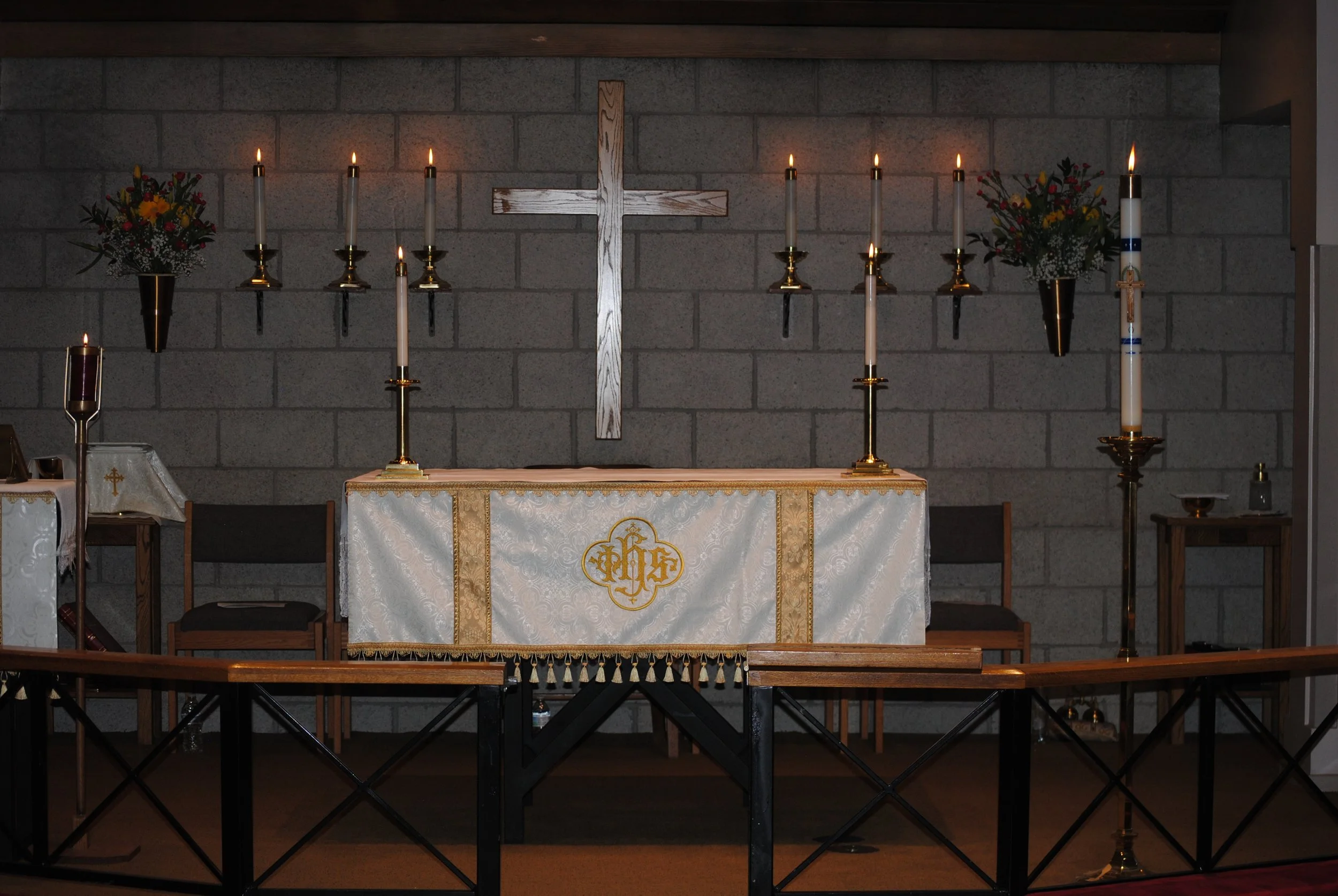 Inside of a church altar with a large wooden cross on a brick wall, lit candles in candleholders, floral arrangements, and a decorated altar cloth with religious symbols.