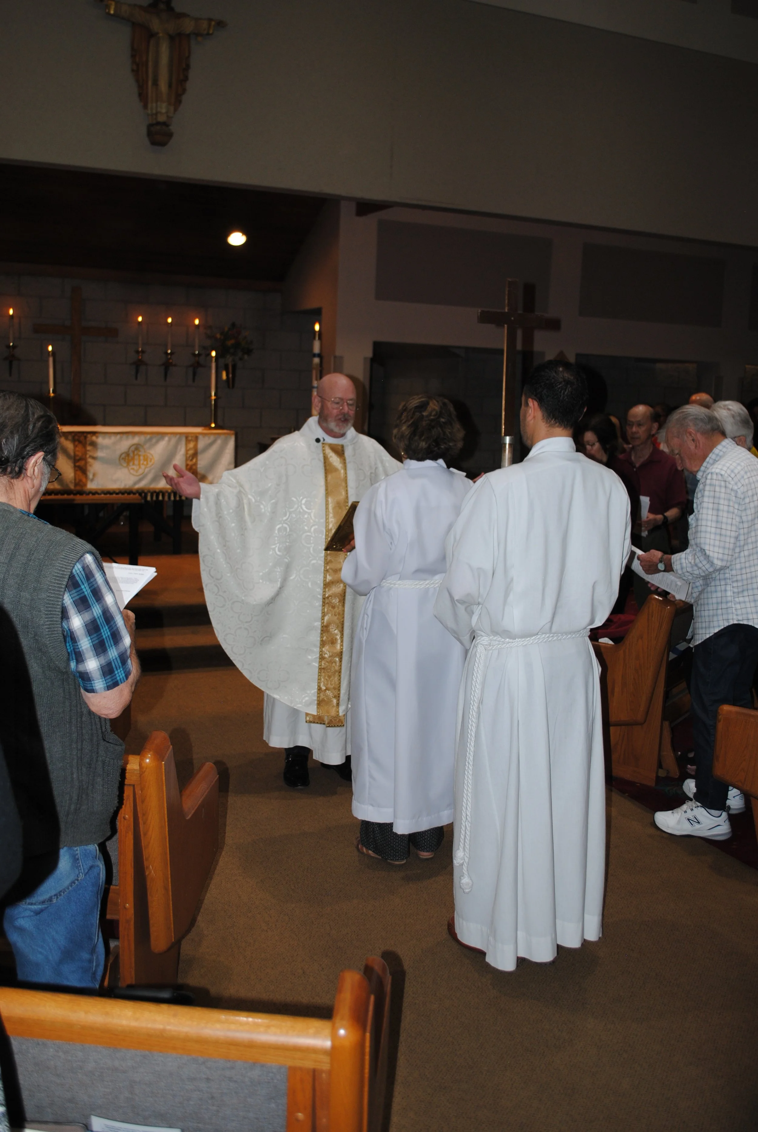A group of people participating in a religious baptism ceremony inside a church. The priest is wearing white and gold vestments, and two individuals are wearing white robes. The congregation is standing and holding prayer books, with candles and cros