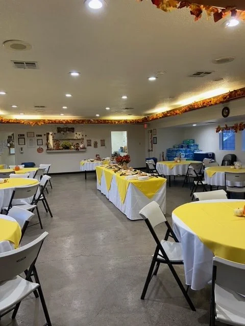 A banquet room decorated for a celebration with round tables covered in white tablecloths and yellow table runners, surrounded by white and black chairs. There are food and drink tables along the walls, with water bottles and snacks. The ceiling has 