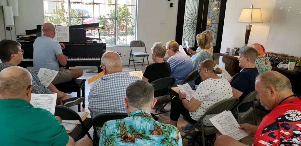 A group of people attending a choir rehearsal or singing class inside a bright room with large windows, seated in chairs, reading music sheets, with a pianist playing at a black grand piano.