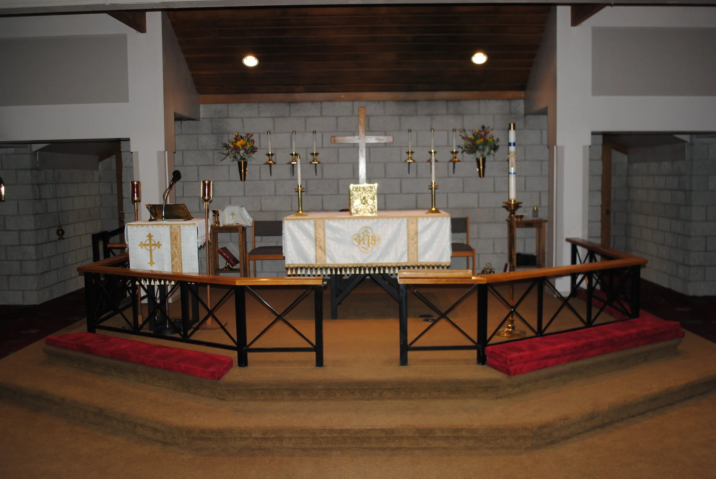 Interior of a church altar with a cross, candles, floral arrangements, and religious items on an altar table, surrounded by a wooden railing.