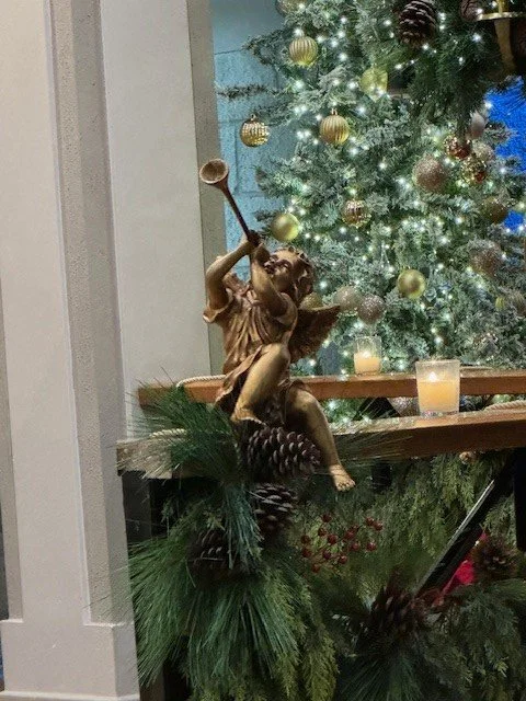 Decorative angel figurine playing a trumpet next to a Christmas tree with ornaments and candles.