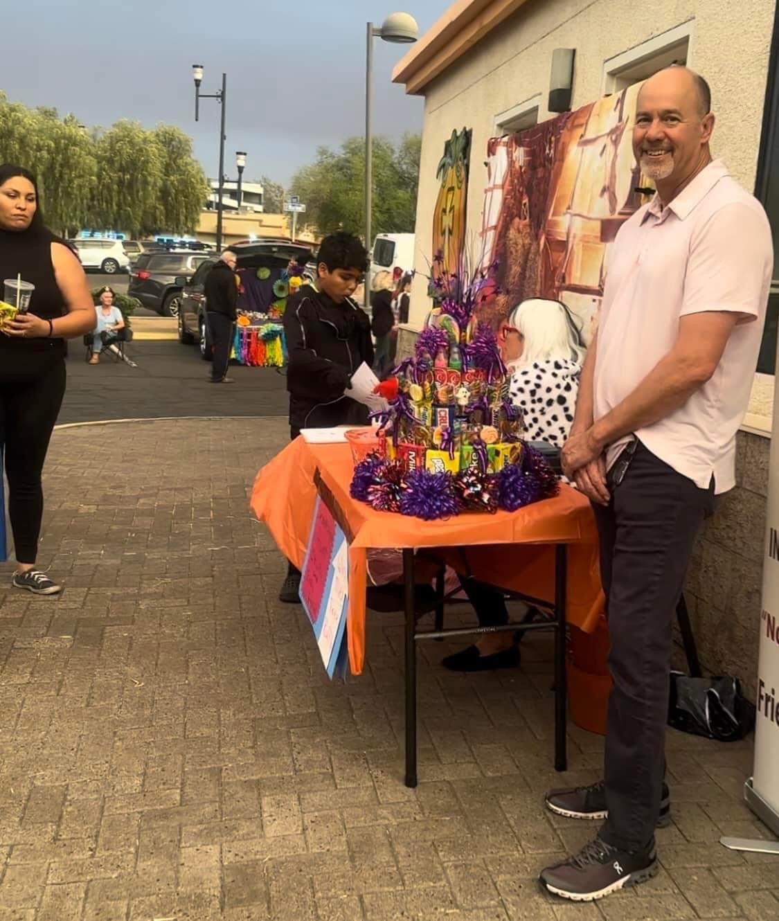 A man standing next to a table decorated with purple and yellow items and candy at an outdoor event. A young boy is looking at the table, and a woman with long dark hair is holding a drink nearby. Several other people are in the background near a har