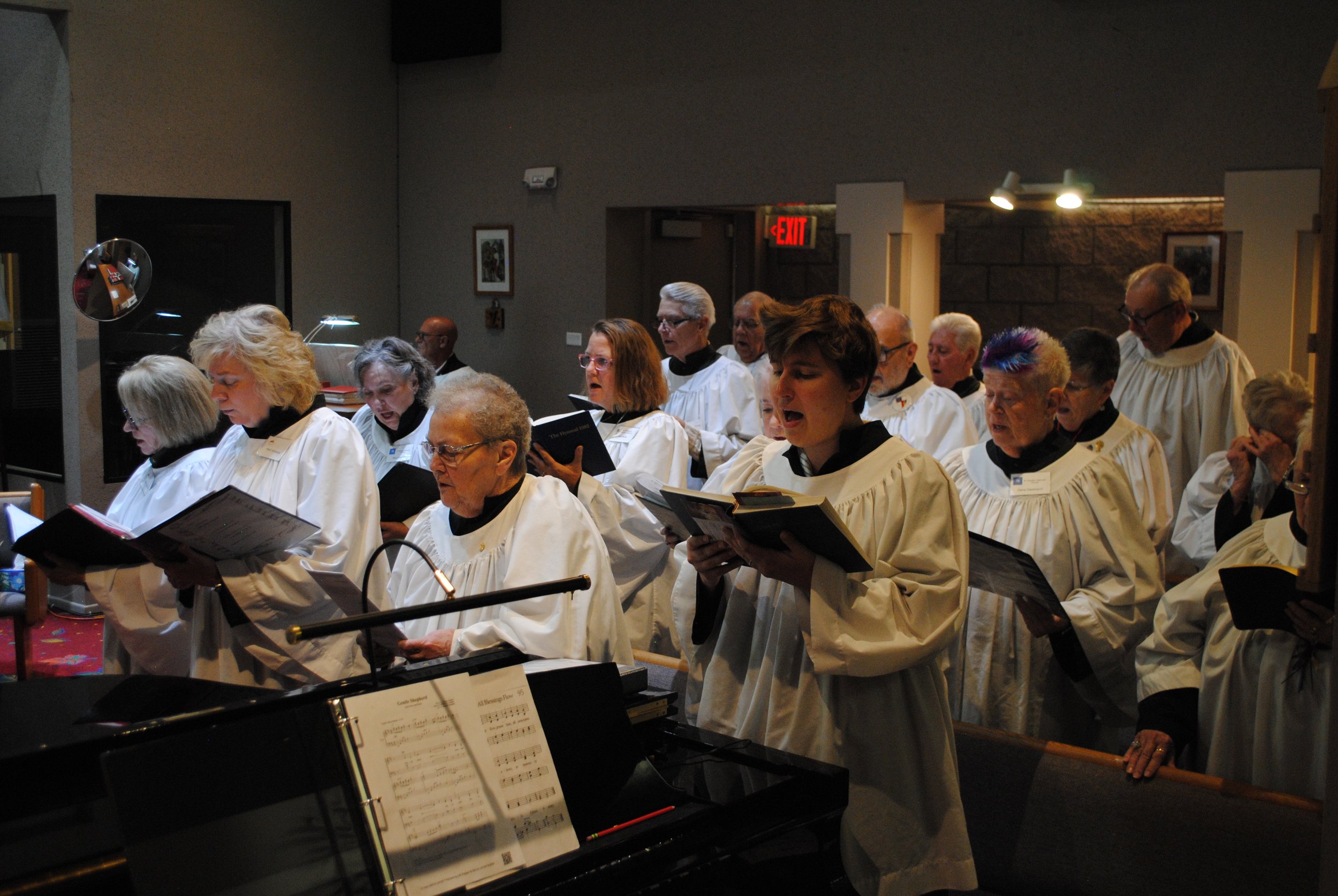 Choir group singing in a church or chapel, led by a conductor, with music sheets and a piano in the foreground.