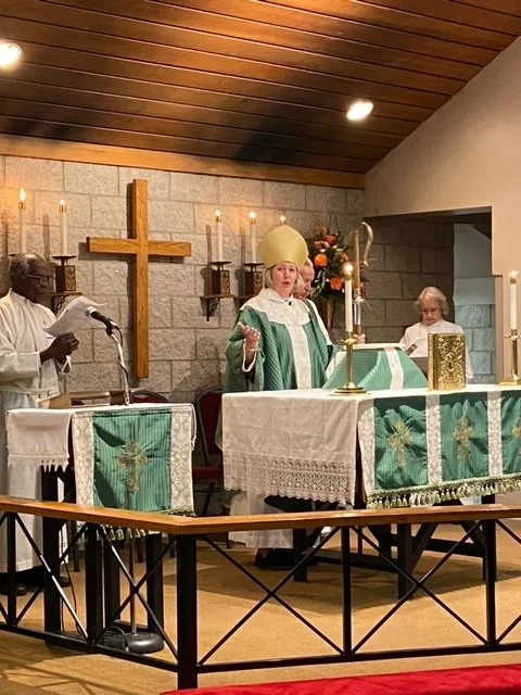 A religious ceremony with a bishop dressed in green and gold vestments, standing behind an altar with a cross and candles. Two other clergy members are present, one reading from a paper and another in white robes. The setting appears to be a church w