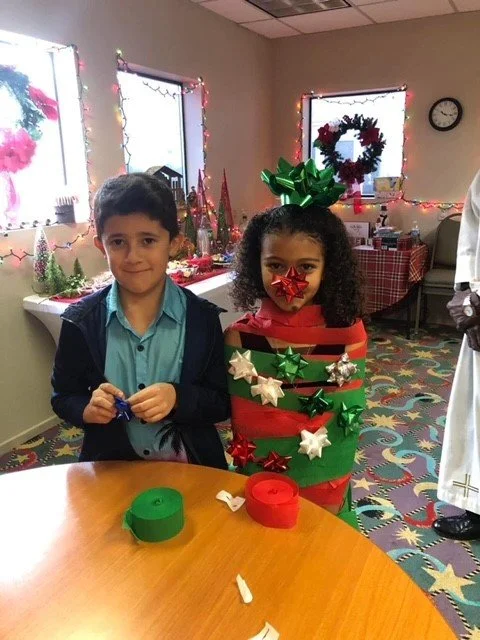 A young boy and girl standing in a decorated room for Christmas. The girl is dressed as a gift box with ribbon and bow decorations, and a large green bow on her head. They are at a table with green and red ribbon rolls. In the background, there are C