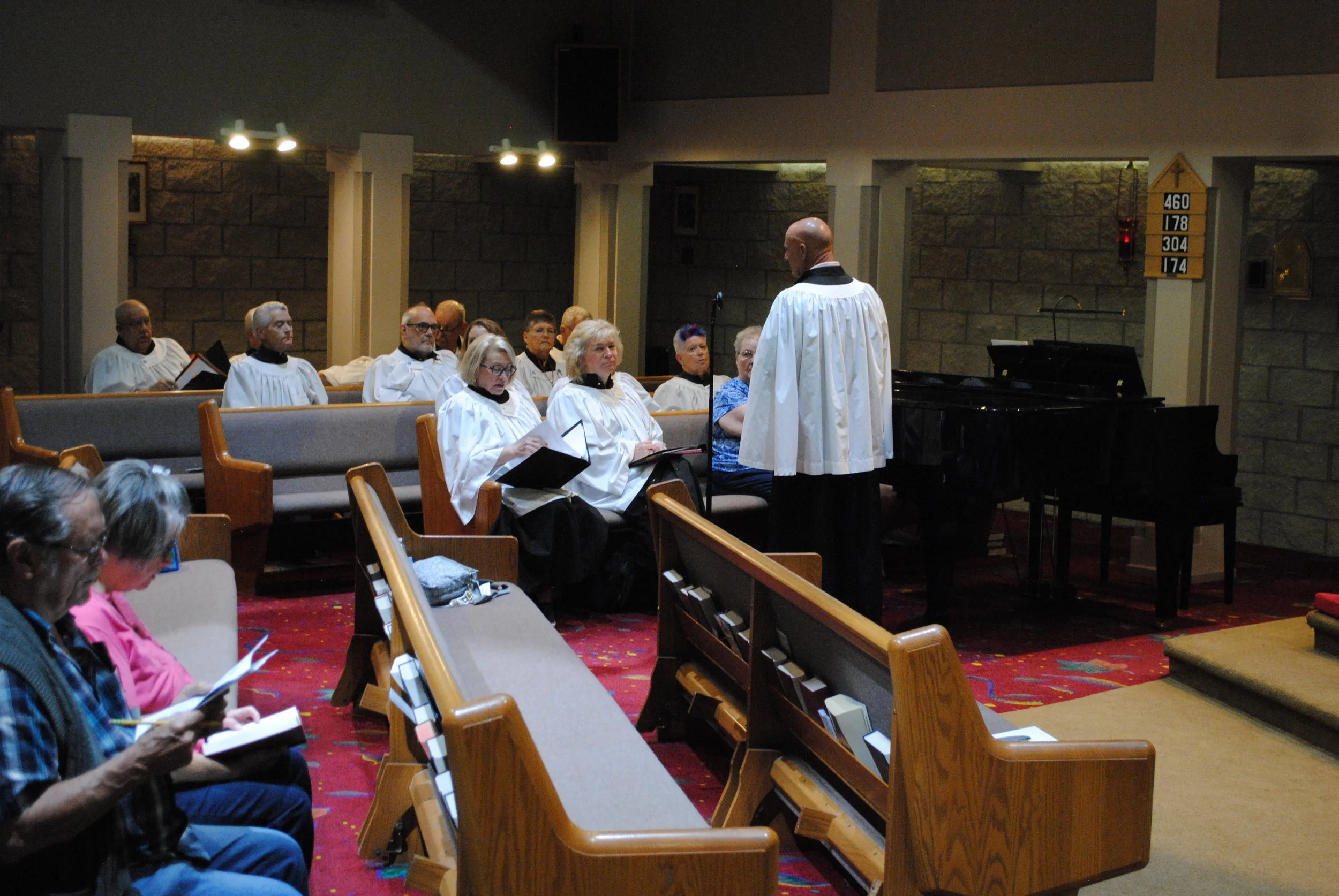 A choir practicing in a church or chapel, with a conductor standing in front of them and a black piano nearby. The choir members are seated on pews and appear to be singing or preparing to sing.