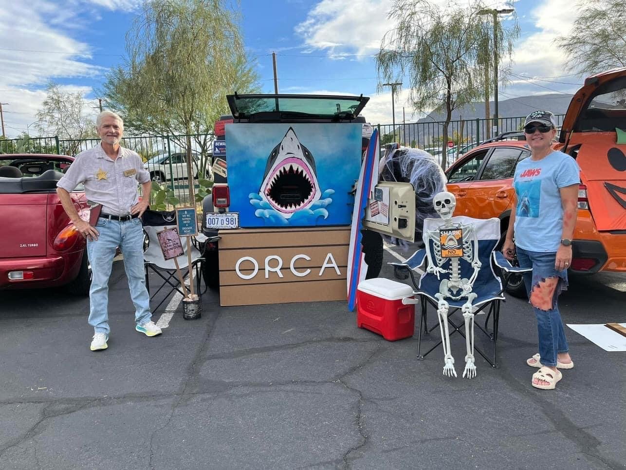 Two people standing in a parking lot next to a mobile shark-themed booth and Halloween decorations, including a skeleton, with cars and trees in the background.