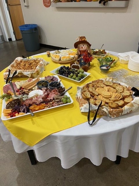 A round table draped with a white tablecloth and yellow overlay holding a variety of snacks and desserts, including grapes, crackers, cheese, cookies, and dips, with a small scarecrow figurine at the center of the table.