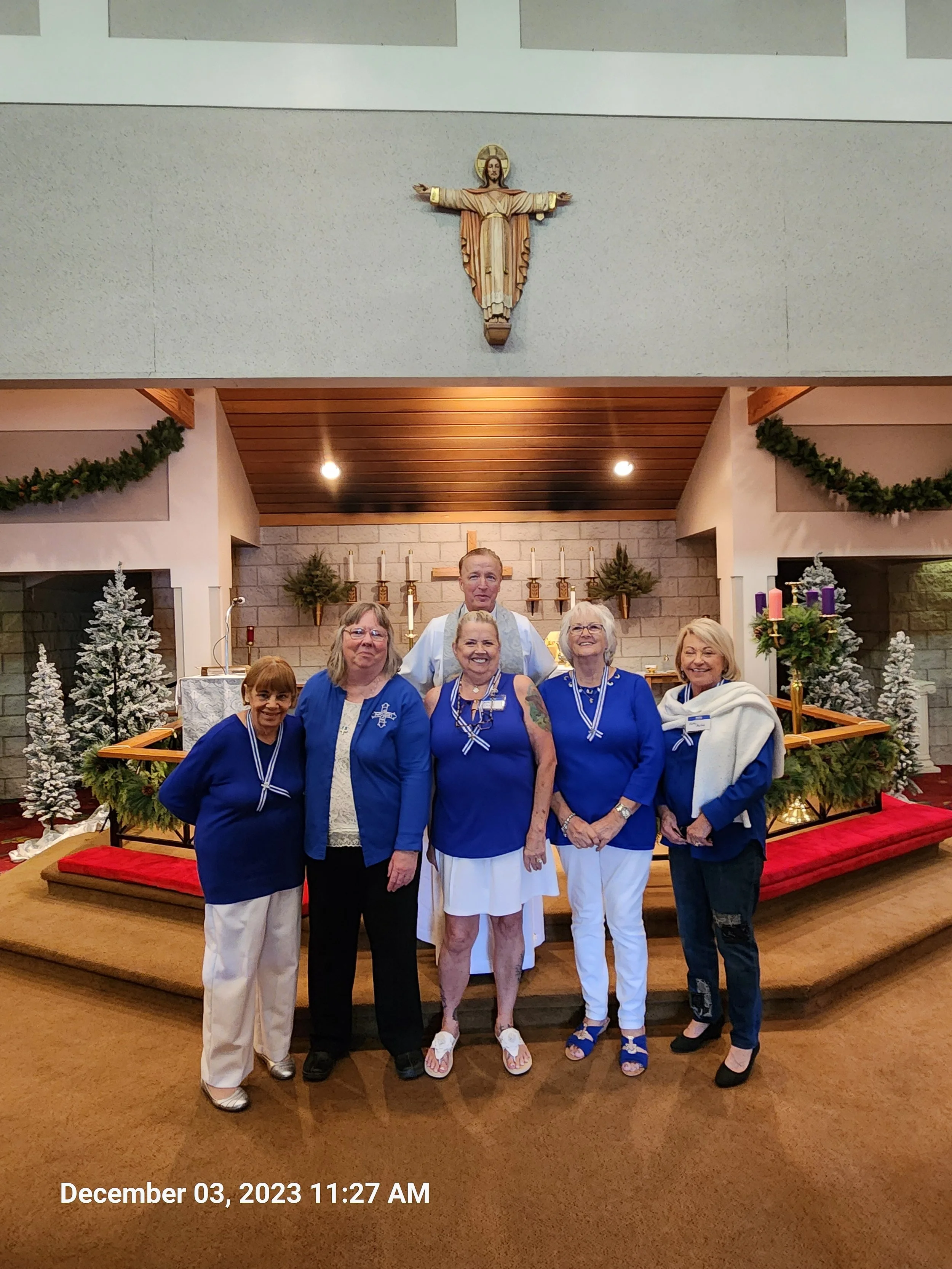 Six women standing in front of an altar inside a church, dressed in blue and white clothing, with Christmas decorations such as small Christmas trees, candles, and garlands around them. A priest is standing behind them.