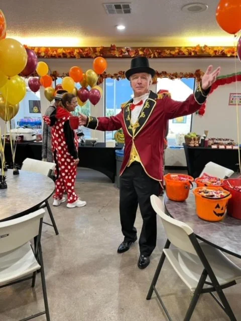 A man dressed as a ringmaster, wearing a red coat with black and gold trim, black top hat, and yellow vest, standing in a decorated room with Halloween-themed orange buckets and balloons, with two women in polka-dot costumes in the background.