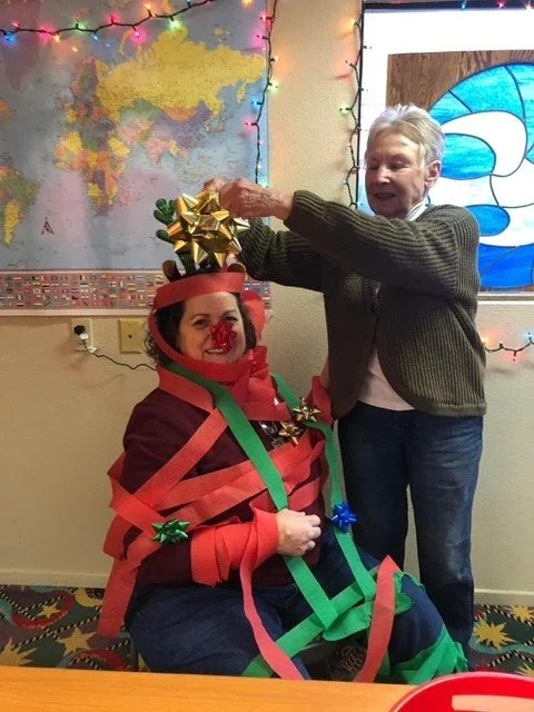 A woman dressed as a Christmas present with red and green ribbons wrapped around her, a red bow on her head, and a red nose, sits on a chair. An older woman, standing beside her, is placing a gold bow on her head. The background shows a world map, co