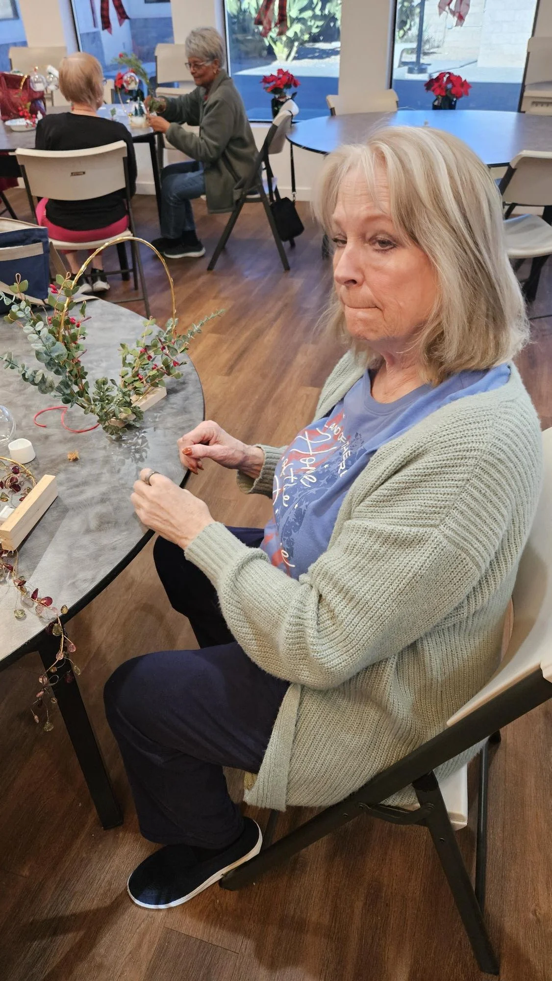 A woman with blonde hair sitting at a table, working on holiday flower arrangements with poinsettias and greenery, in a room decorated for Christmas.