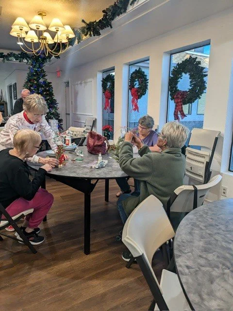 Four elderly women decorating Christmas ornaments at a table in a festive room with Christmas wreaths, garlands, and a decorated Christmas tree.