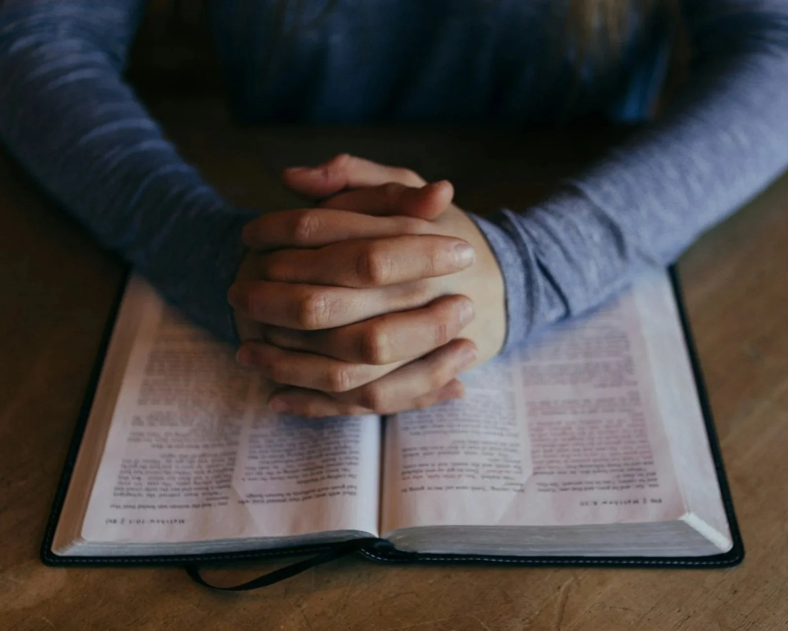 Person praying with hands clasped over an open Bible on a wooden table.