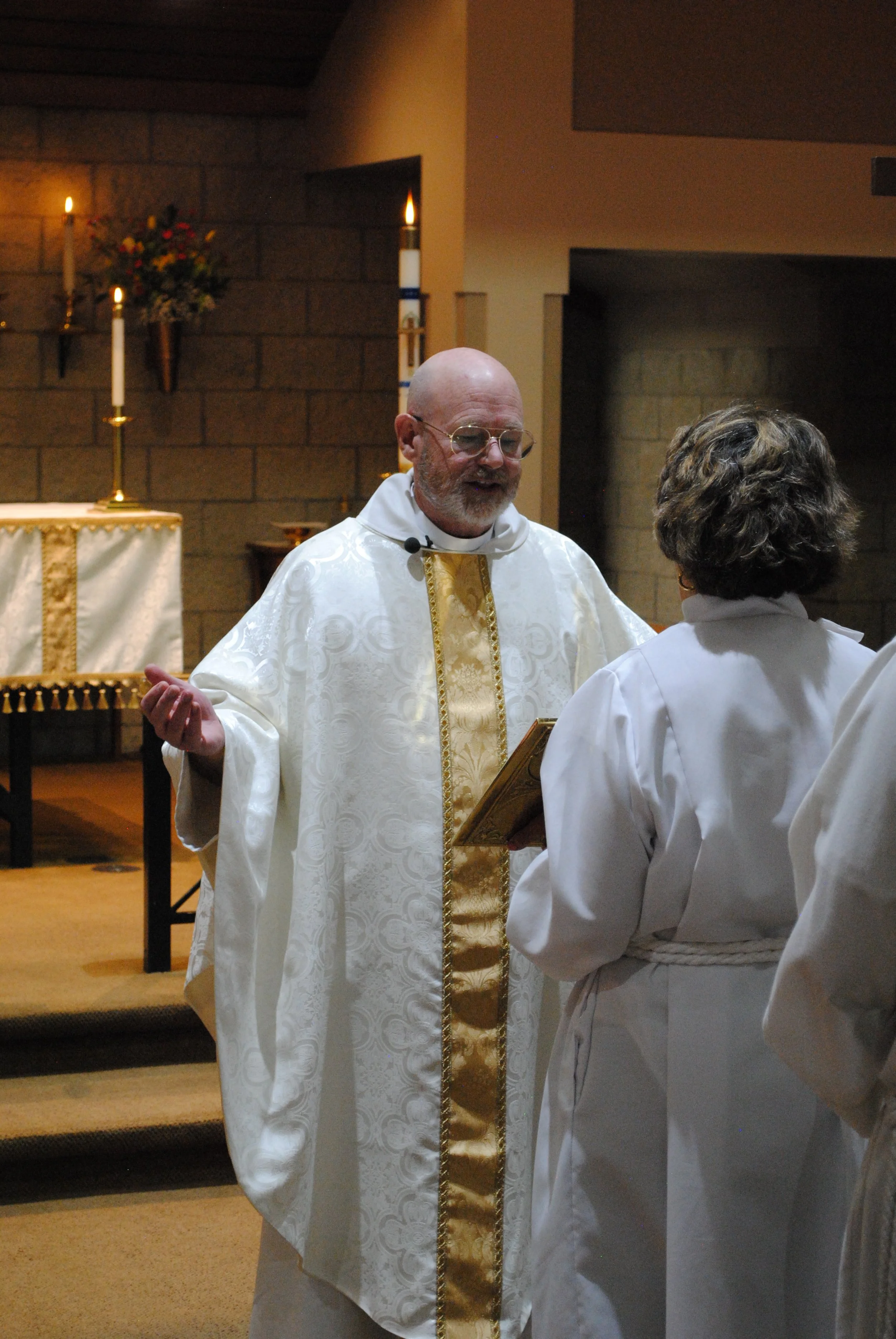 A priest wearing white and gold liturgical vestments is conducting a religious ceremony inside a church, with a woman in white robes standing in front of him.