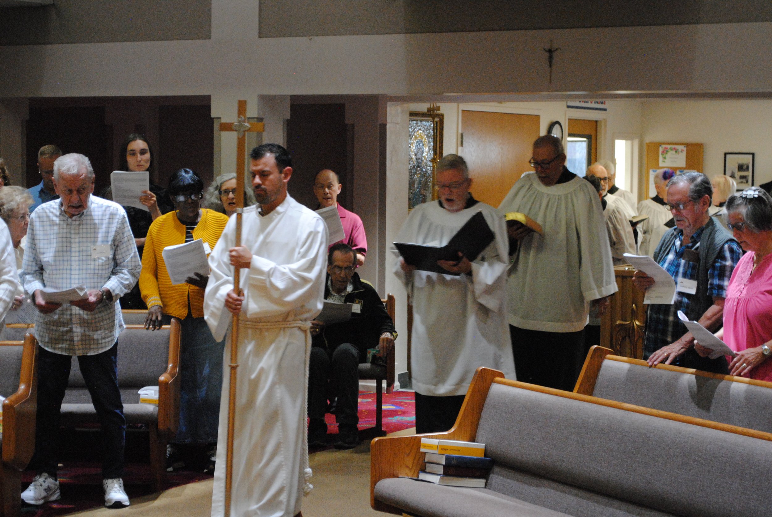People gathered in a church during a religious service, some holding or reading from hymnals or prayer books, with a priest or minister in white robes leading the congregation.