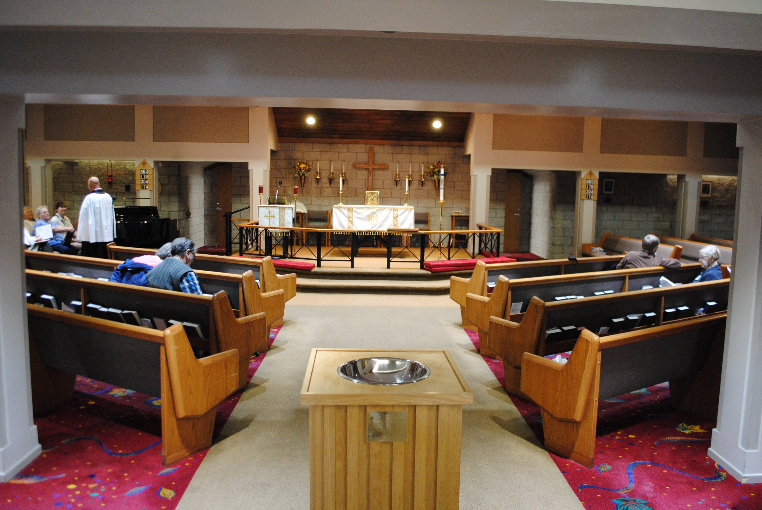 Interior view of a church altar with wooden pews on either side, a small red carpet in front, and an altar with candles, a cross, and floral arrangements. Several elderly people are seated in the pews, and a priest or minister is standing near the gr