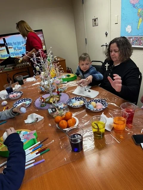 Easter celebration with children and an adult at a table filled with decorated eggs, juice, and a colorful centerpiece. A woman with curly hair and a child are sitting at the table, and another woman is in the background watching a screen.
