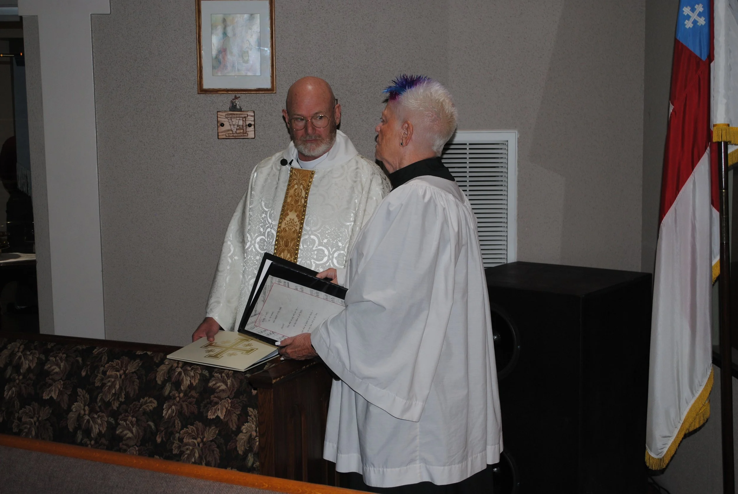 A religious ceremony with clergy members, one holding a framed document, inside a church with framed art and an American flag in the background.