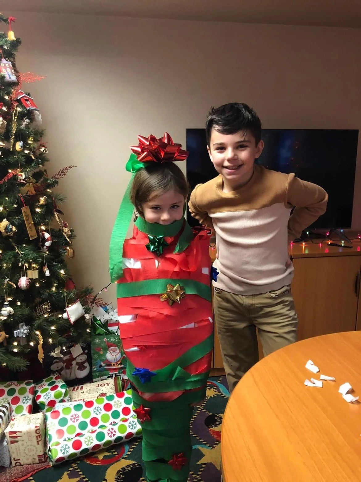 A young girl dressed as a Christmas present with red, green, and gold decorations, standing next to a boy in a beige and brown sweater, both smiling in a room decorated for Christmas with a decorated tree and wrapped gifts.