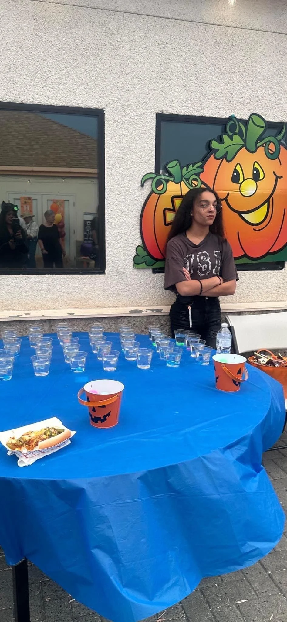 A girl stands with arms crossed in front of a pumpkin-themed decoration at a Halloween party, with a table of cups and snacks nearby.