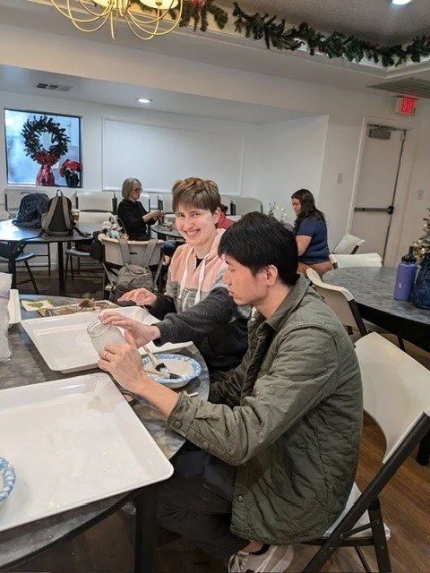 Two people sit at a festive holiday table with empty trays, smiling and engaging with each other. In the background, others are seated, and there is holiday decor including a wreath and garland, with a window showing a holiday scene.