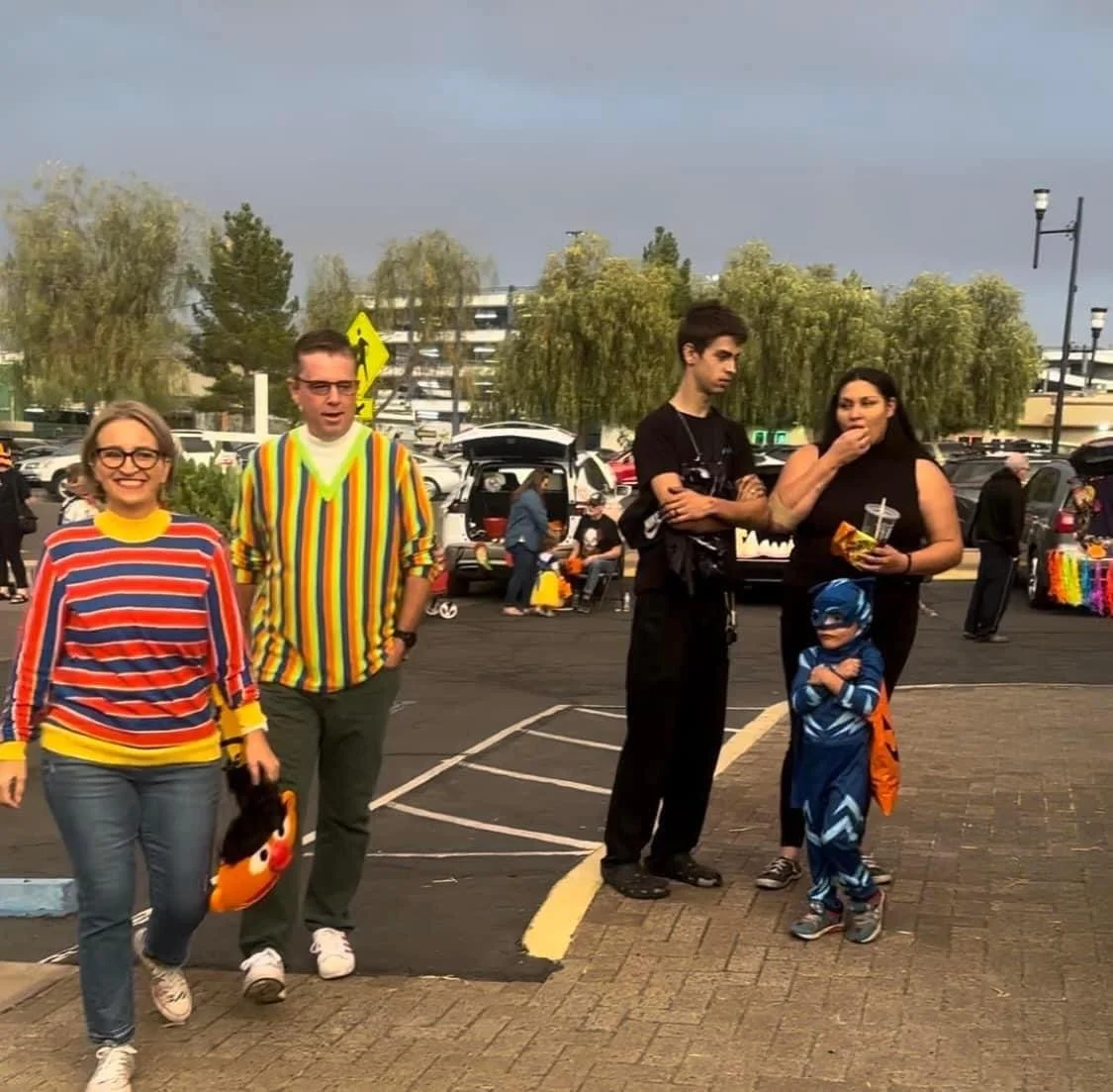 Group of five people standing in a parking lot on a cloudy day, some dressed in comic book and rainbow costumes, with cars and trees in the background.